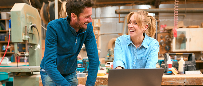 a male and a female carpenter sharing a laugh while working on a laptop in thier wood working shop.